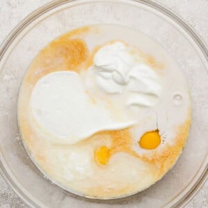 A glass mixing bowl containing an egg, milk, yogurt, and other liquid baking ingredients on a light textured surface.