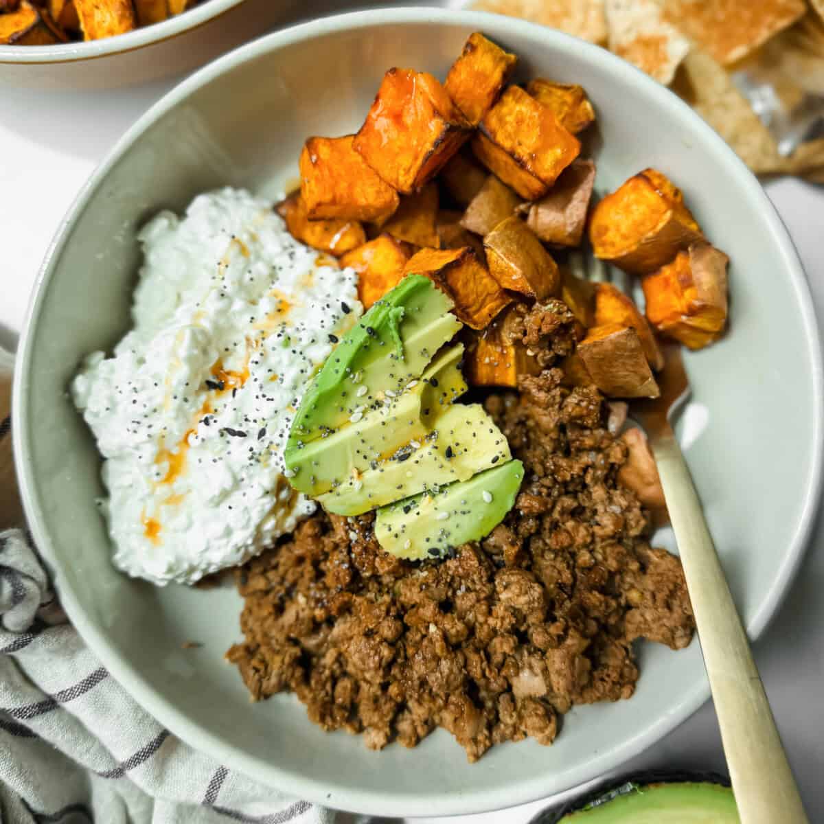 A bowl containing ground beef, roasted sweet potatoes, cottage cheese with hot sauce, and avocado slices, topped with black pepper.