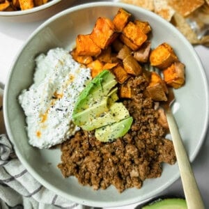 A bowl containing ground beef, roasted sweet potatoes, cottage cheese with hot sauce, and avocado slices, topped with black pepper.