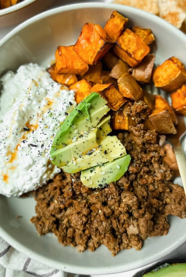 A bowl containing ground beef, roasted sweet potatoes, cottage cheese with hot sauce, and avocado slices, topped with black pepper.