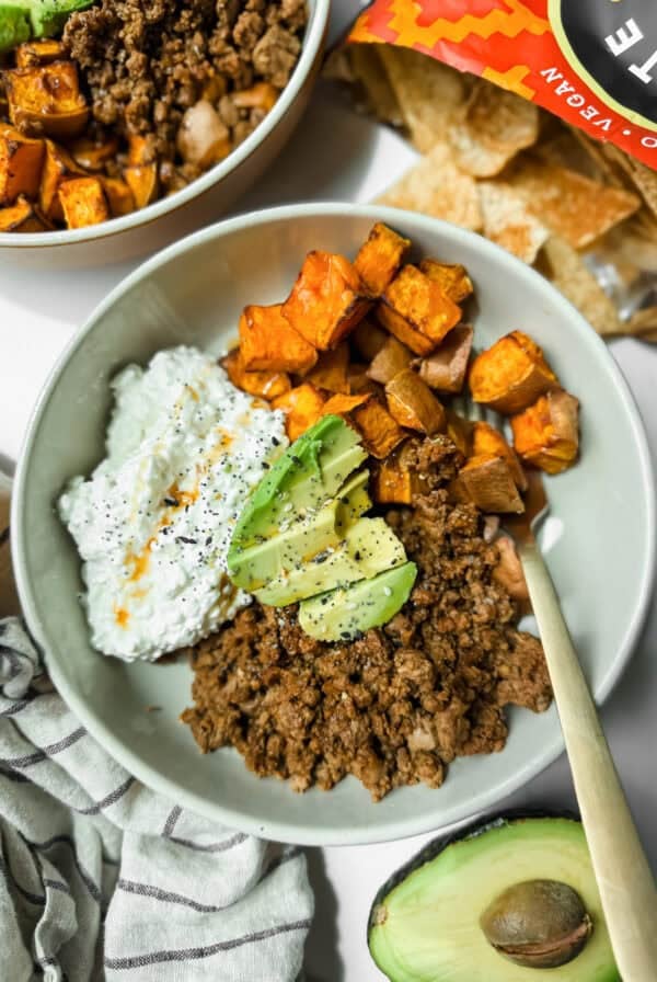 A bowl with ground beef, roasted sweet potatoes, cottage cheese, and avocado slices sits next to an open bag of tortilla chips and a halved avocado.