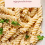 A bowl of spiral pasta in creamy cheese sauce, garnished with parsley and bread crumbs. Text overlay reads "Cottage Cheese Mac & Cheese. High-protein dinner!.