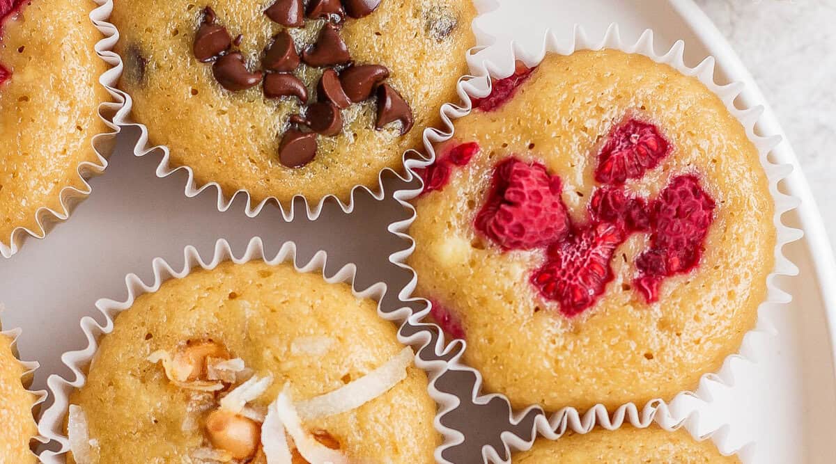 Three muffins on a white plate, each topped with chocolate chips, chopped raspberries, and coconut shreds—a perfect treat for fit foodie finds enthusiasts.