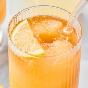 A close-up of an iced orange drink in a ribbed glass with a clear straw and a lemon slice garnish.