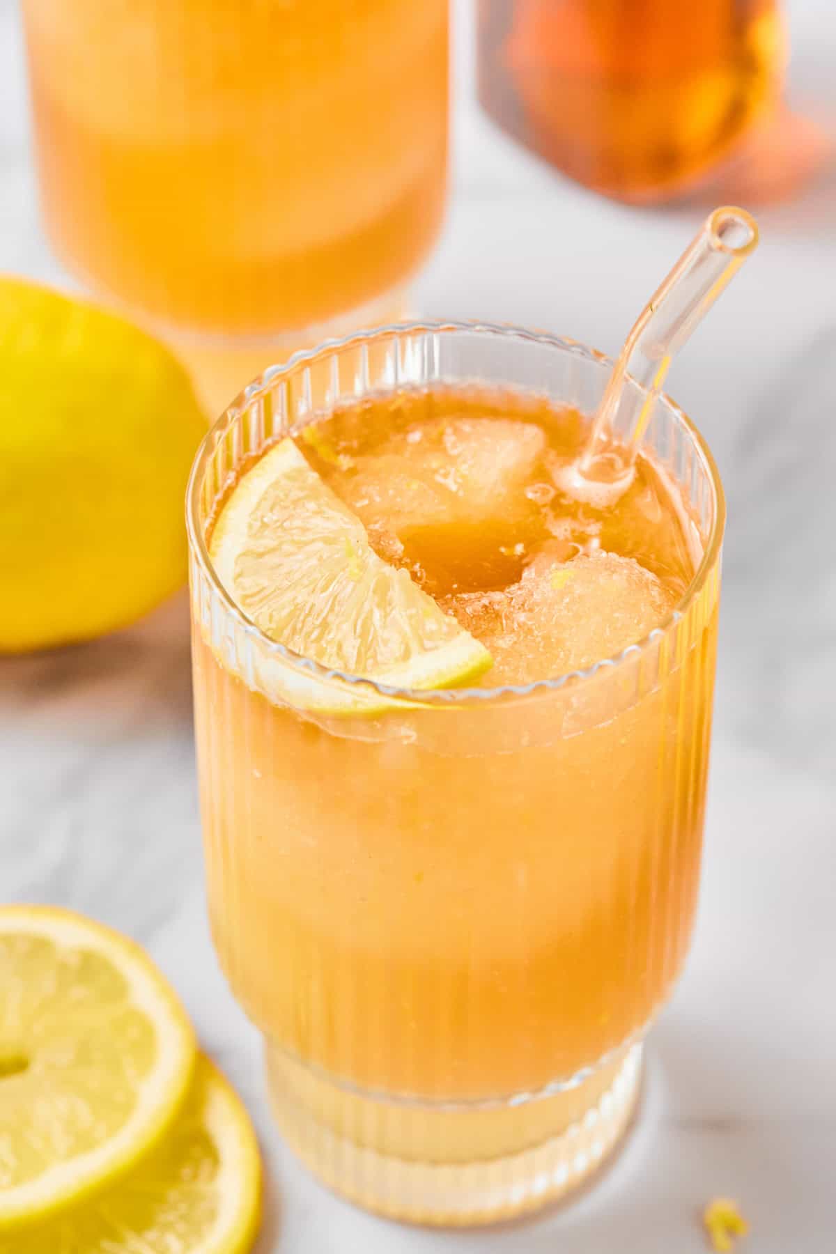 A glass of iced lemon drink with a slice of lemon on the rim and a glass straw, placed on a light surface with lemon slices nearby.