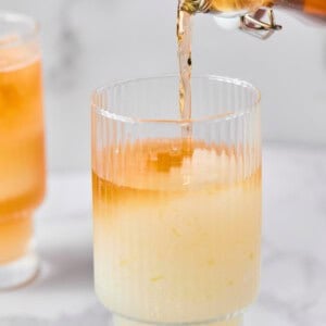 A glass with a pale yellow liquid and ice is being filled with an orange liquid from a glass bottle. Another similar drink is in the background.