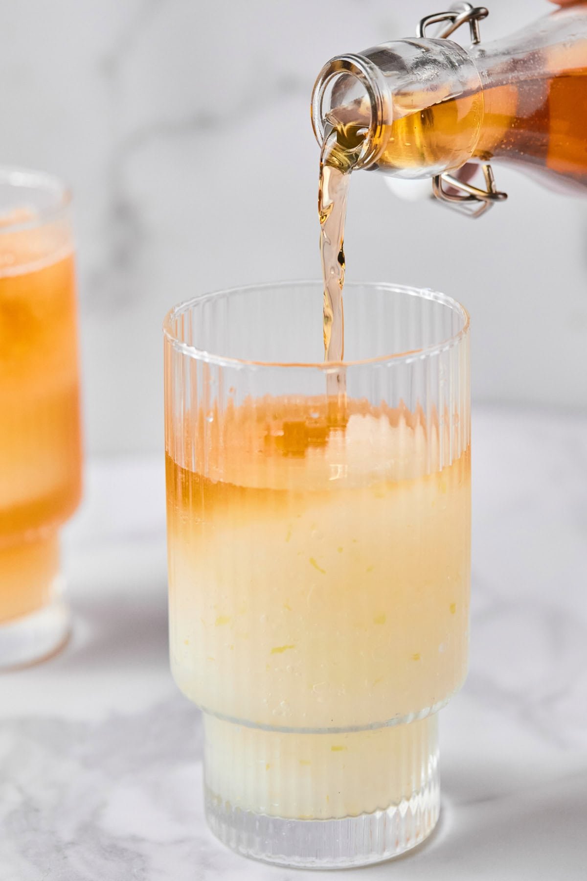 A glass with a pale yellow liquid and ice is being filled with an orange liquid from a glass bottle. Another similar drink is in the background.