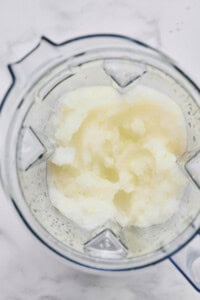 A top-down view of a blender containing a slushy, pale mixture with a slightly grainy texture on a white surface.