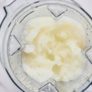 A top-down view of a blender containing a slushy, pale mixture with a slightly grainy texture on a white surface.