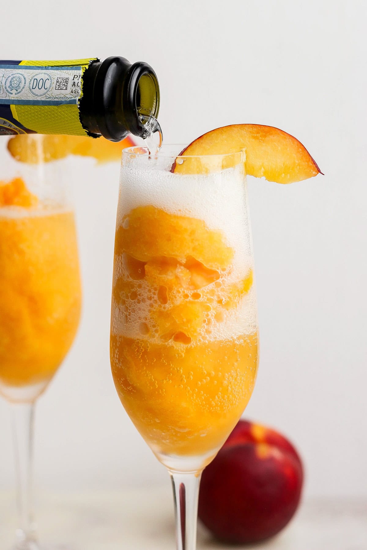 A glass filled with peach sorbet is being topped with sparkling wine, garnished with a peach slice; another glass and a peach are in the background.