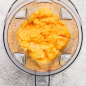 Top-down view of an open blender containing a smooth, orange-yellow mixture on a light textured surface.