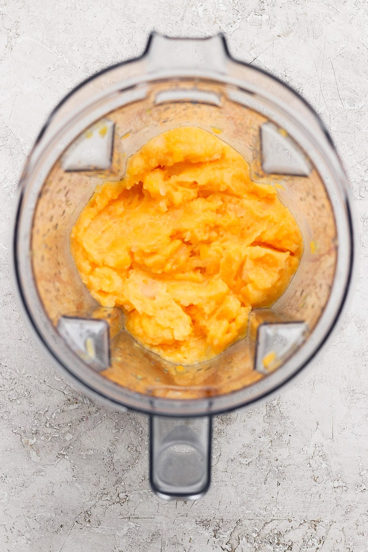 Top-down view of an open blender containing a smooth, orange-yellow mixture on a light textured surface.