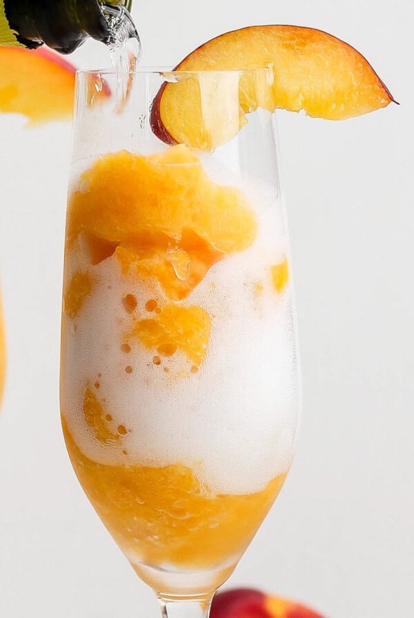 A champagne flute filled with orange sorbet and topped with bubbly drink, garnished with a peach slice on the rim.