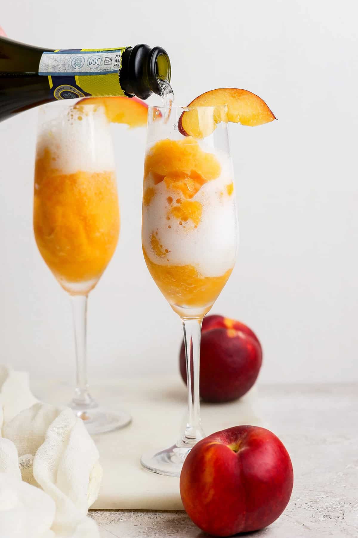 A glass of peach sorbet with sparkling wine being poured over it, garnished with a peach slice; another filled glass and whole peaches are in the background.