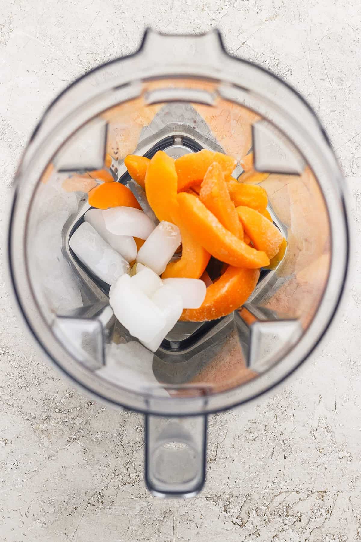 Top-down view of a blender containing ice cubes and orange fruit slices on a light-colored surface.