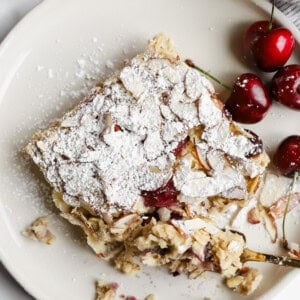 A square slice of almond cherry cake topped with powdered sugar on a plate, served with fresh cherries on the side.