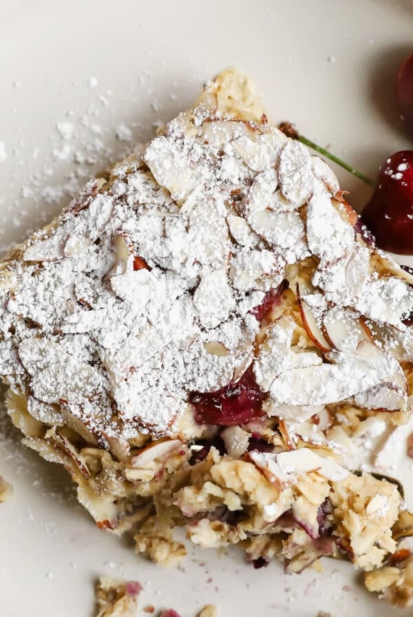 A square slice of almond cherry cake topped with powdered sugar on a plate, served with fresh cherries on the side.