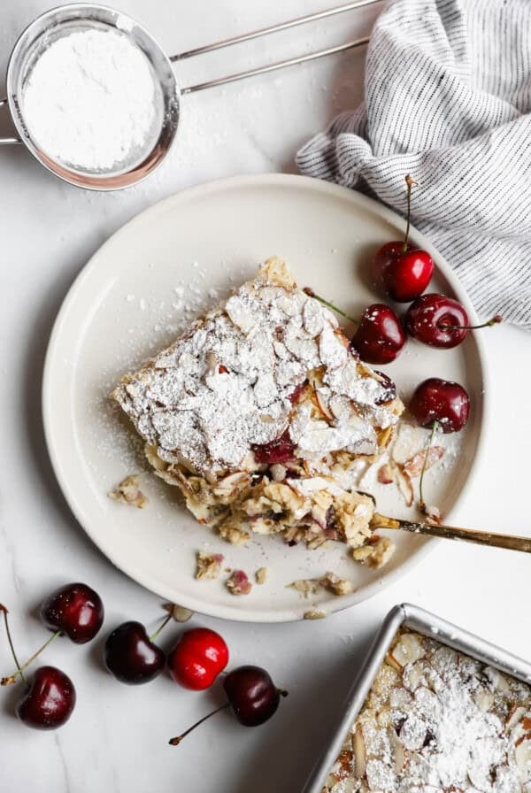 A slice of cherry dessert dusted with powdered sugar on a plate, surrounded by fresh cherries, a striped cloth, and a sifter with powdered sugar.