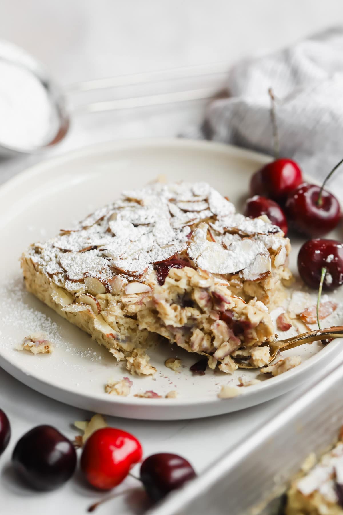 A partially eaten slice of almond and cherry cake topped with powdered sugar on a plate, with fresh cherries on the side.