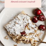 A plate with a slice of cherry almond baked oatmeal topped with powdered sugar and almond flakes, surrounded by fresh cherries, on a white background.