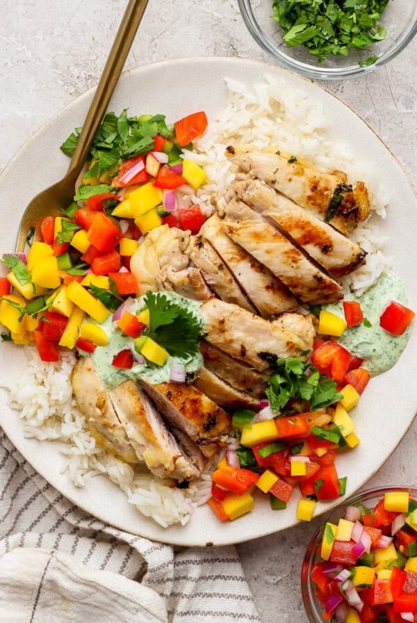 A plate of sliced grilled chicken breast served over white rice with chopped mango, red bell pepper, red onion, herbs, and a dollop of green sauce.