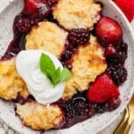 A bowl of mixed berry cobbler with blackberries and strawberries, topped with a dollop of whipped cream and a mint leaf.