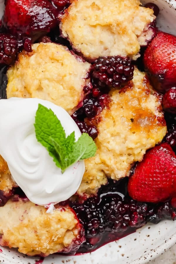 A bowl of mixed berry cobbler with blackberries and strawberries, topped with a dollop of whipped cream and a mint leaf.