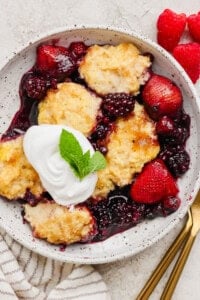 A bowl of mixed berry cobbler topped with whipped cream and a mint leaf, served with fresh strawberries and raspberries on the side.