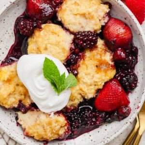 A bowl of mixed berry cobbler topped with whipped cream and a mint leaf, served with fresh strawberries and raspberries on the side.