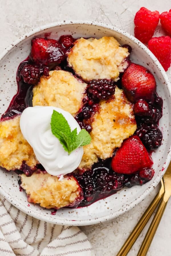 A bowl of mixed berry cobbler topped with whipped cream and a mint leaf, served with fresh strawberries and raspberries on the side.