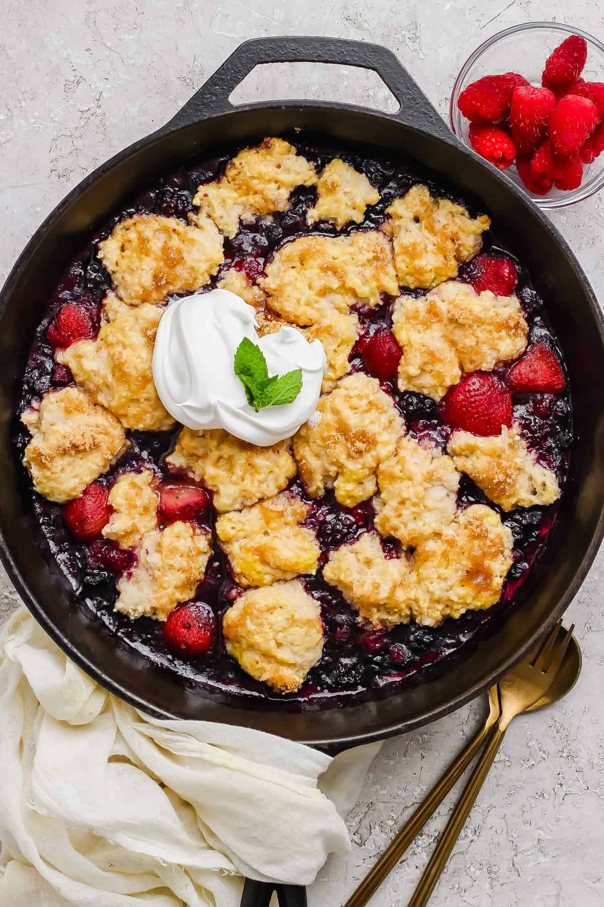 A skillet of mixed berry cobbler topped with golden biscuit dough, a dollop of whipped cream, a mint garnish, and a side of fresh raspberries.
