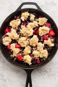A cast iron skillet filled with mixed berries topped with dollops of crumbly dough and brown sugar, ready for baking.