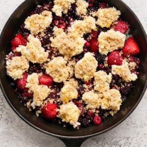 A cast iron skillet filled with mixed berries topped with dollops of crumbly dough and brown sugar, ready for baking.