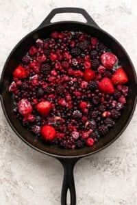 A cast iron skillet filled with a mix of frozen berries, including strawberries, raspberries, blackberries, and blueberries, on a light textured surface.