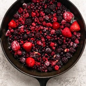 A cast iron skillet filled with a mix of frozen berries, including strawberries, raspberries, blackberries, and blueberries, on a light textured surface.