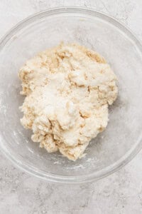 A glass bowl containing a rough, partially-mixed dough on a light-colored surface.