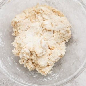A glass bowl containing a rough, partially-mixed dough on a light-colored surface.