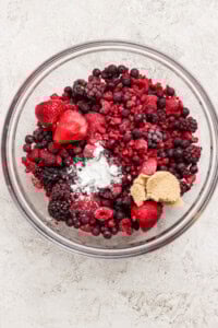 A glass bowl filled with mixed frozen berries, a mound of brown sugar, and a small amount of white powder on a light surface.