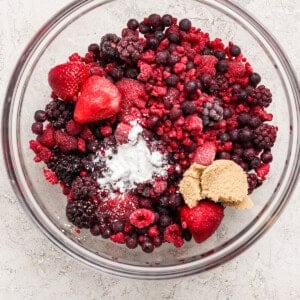 A glass bowl filled with mixed frozen berries, a mound of brown sugar, and a small amount of white powder on a light surface.