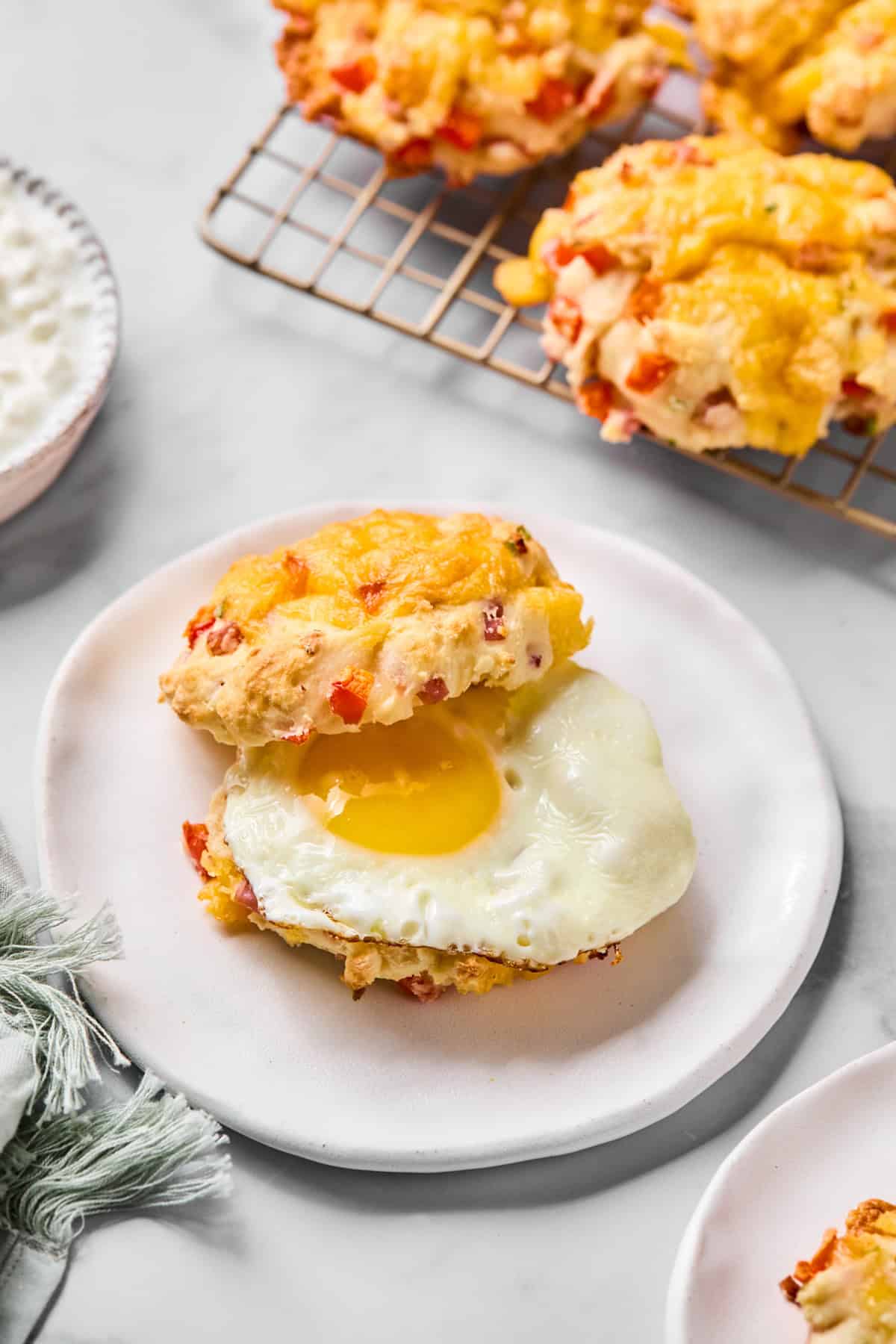 A breakfast sandwich with a fried egg inside a cheddar and red pepper biscuit, served on a white plate, with more biscuits on a cooling rack in the background.
