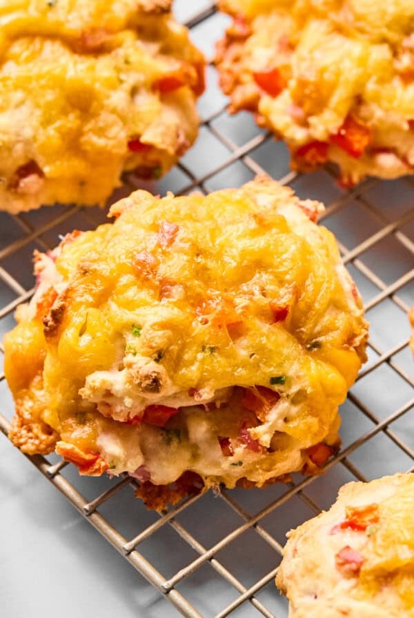 Six cheesy baked biscuits with visible red pepper pieces cooling on a wire rack against a light background.