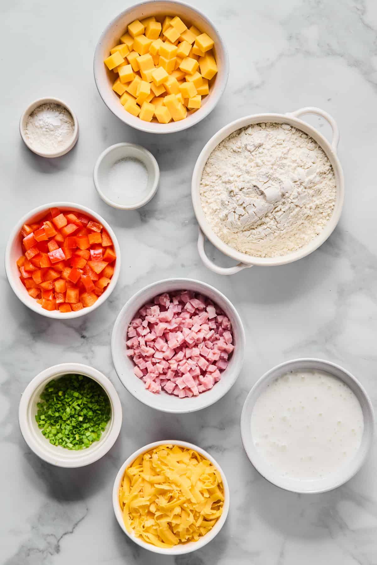 Assorted ingredients in bowls on a marble surface, including cubed cheddar cheese, flour, milk, diced ham, diced red bell pepper, grated cheese, chopped chives, salt, and baking powder.