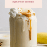 A glass of peanut butter banana smoothie topped with whipped cream and peanut butter, with a banana and another glass in the background.