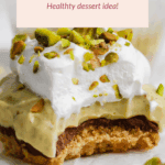 A pistachio cheesecake bite topped with whipped cream and chopped pistachios, resting on a crumbly base, with a bite taken out to show the layers.