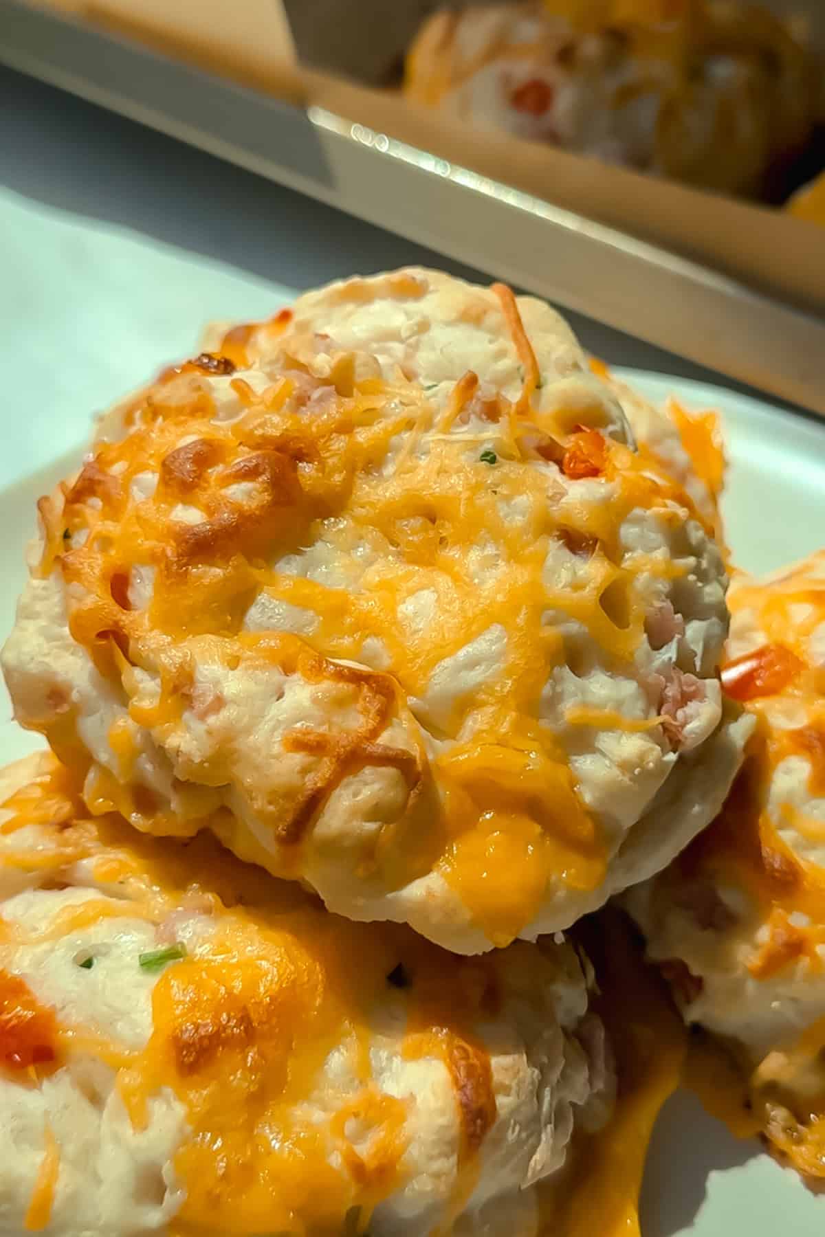 Close-up of two cheesy biscuits with melted cheddar on top, placed on a white plate with more biscuits blurred in the background.