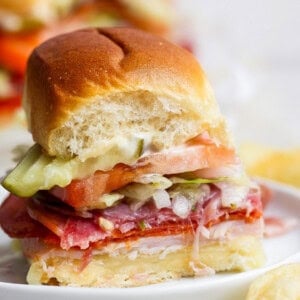 A small sandwich with sliced meats, cheese, tomato, lettuce, pickles, and mayonnaise on a soft bun, shown with a bite taken out of it.