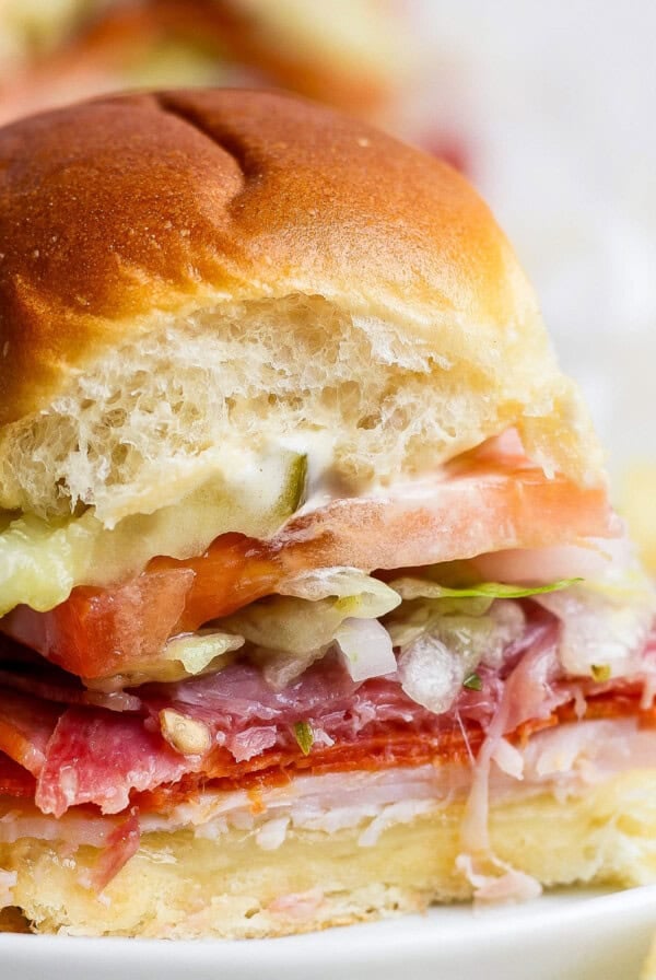 A small sandwich with sliced meats, cheese, tomato, lettuce, pickles, and mayonnaise on a soft bun, shown with a bite taken out of it.