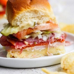A small sandwich on a white plate, layered with deli meats, cheese, lettuce, tomato, and pickles, with a bite taken out. Potato chips are visible beside the plate.