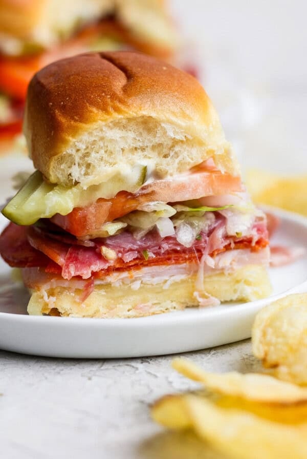 A small sandwich on a white plate, layered with deli meats, cheese, lettuce, tomato, and pickles, with a bite taken out. Potato chips are visible beside the plate.