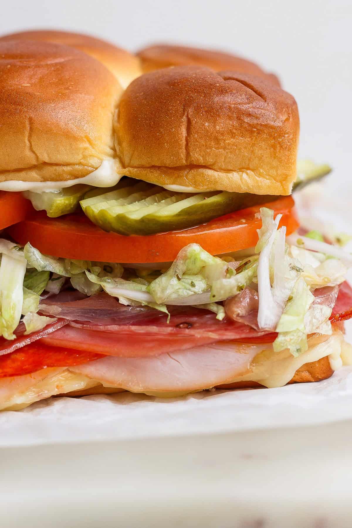 A close-up of a sandwich with sliced meats, shredded lettuce, tomato, pickles, mayonnaise, and a soft, browned bun.
